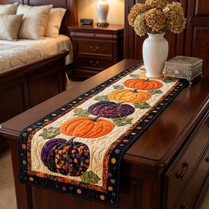 May include: A rectangular quilted table runner featuring a repeating pattern of orange, yellow, and purple pumpkins with green leaves on a cream background, framed by a black border with colorful polka dots and a decorative stitched texture.