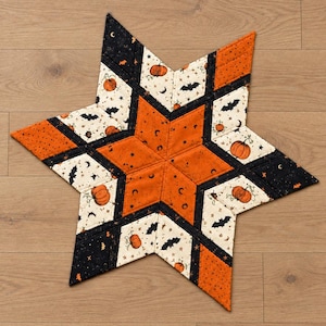 May include: A star-shaped quilted fabric mat featuring a patchwork design. The pattern includes orange fabric with black stars, cream fabric with orange pumpkins and black bats, and solid black fabric with small orange speckles throughout the design.