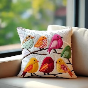 May include: A decorative white pillow featuring appliqued birds in various colors. The birds are perched on brown branches, with details including orange, pink, yellow, and green. The pillow has a lace-like trim.