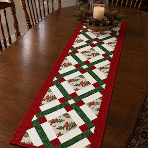 May include: A rectangular table runner featuring a patchwork design with a red border. The center pattern consists of white squares with a cardinal bird and pine branch print, interconnected by dark green and red fabric strips in a geometric layout.