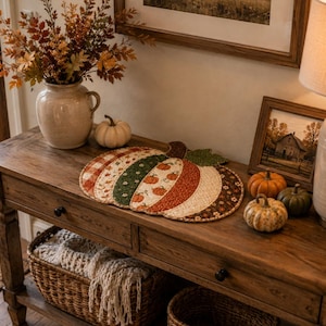 May include: A quilted fabric placemat shaped like a pumpkin, featuring alternating vertical panels of various autumn-themed patterns including orange plaid, green floral, and pumpkin prints, finished with a green fabric leaf at the top stem.