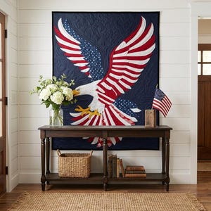 May include: A rectangular wall quilt featuring a large, central bald eagle with wings spread wide. The eagle is designed with red and white stripes and blue sections with white stars, mimicking the American flag. The background is dark navy blue.