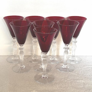 May include: A set of eight ruby red wine glasses. Each glass features a clear, ornate stem and a wide, conical bowl. The glasses are arranged on a light-colored surface, showcasing their elegant design and vibrant color.