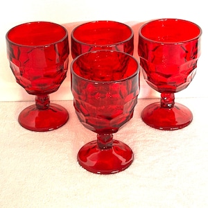 May include: Four ruby red goblets with a faceted design. The stemware has a round base, a short stem, and a wide bowl. The glasses are made of a transparent material, allowing light to pass through. The goblets are arranged on a white surface.