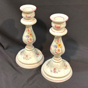 May include: Two white ceramic candlesticks with floral designs. Each candlestick has a wide base, a central stem, and a top cup for holding a candle. The floral pattern includes pink, yellow, and blue flowers, with red trim.