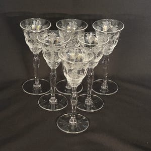 May include: Six clear crystal wine glasses with etched floral designs. The glasses have a slender stem and a round base. The etched design is visible on the bowl of the glass. The glasses are arranged in a circular pattern.