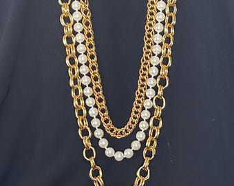 May include: A layered necklace featuring gold-tone chains and strands of faux pearls. The necklace has three distinct layers, with varying chain styles and pearl sizes. The background is a solid dark color.