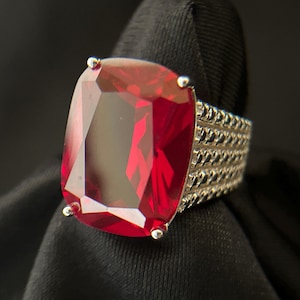 May include: A large, rectangular red gemstone ring with a silver band. The gemstone is faceted, and the band features a textured design. The ring is set against a dark background, highlighting its vibrant color and intricate details.