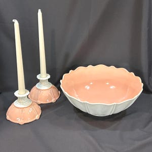May include: A decorative set featuring two pink and white candle holders with cream-colored candles and a large, scalloped-edge bowl. The bowl has a pink interior and a white exterior.