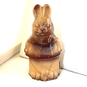 May include: A brown and tan bunny figurine with a glossy finish. The bunny is in a seated position with its paws resting on its lap. The figurine has large ears and a detailed face. The base of the figurine is a cylindrical shape.