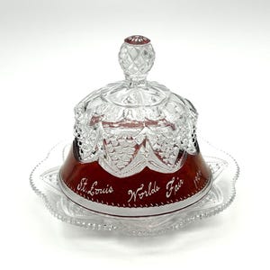May include: A clear and deep red glass butter dish with a matching lid. The red glass section features the etched text St. Louis Worlds Fair 1904. The lid has a faceted knob and a clear glass top with a textured pattern, resting on a clear glass plate.