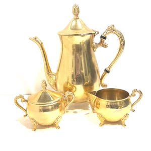 May include: A gold-colored tea set including a teapot, sugar bowl, and creamer. The teapot has a curved spout and ornate handle. The sugar bowl has a lid and handles. The creamer has a small spout and handle. All pieces have decorative feet.