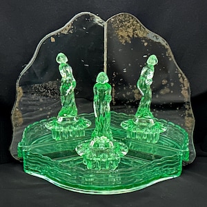 May include: A green glass centerpiece with three figures and a decorative tray. The figures are standing and have a flowing design. The tray has a wavy edge and is made of the same green glass. The background is a mirrored surface.