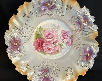 May include: A decorative porcelain plate with a ruffled edge. The plate features a central floral design of pink roses and surrounding embossed flowers with gold accents. The rim has a peach-colored border.
