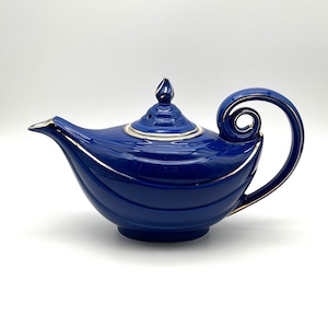 May include: A deep blue ceramic teapot with a gold-trimmed rim, spout, and lid. The teapot features a curved, swirling handle and a pointed finial on the lid. The body has a smooth, rounded shape with subtle horizontal ridges and gold accents.