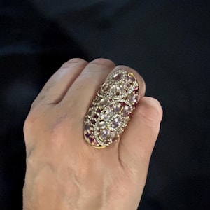 May include: An ornate gold-toned ring featuring an oval design. The ring is encrusted with clear and purple gemstones, accented with small white stones. The ring's design is elongated and covers a significant portion of the finger.