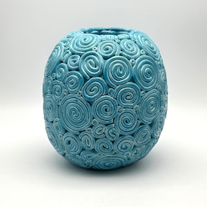 May include: A round, light blue ceramic vase featuring a textured surface covered in numerous raised, coiled spiral patterns of varying sizes. The vessel has a small circular opening at the top and a glossy finish that reflects light across the curves.
