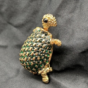 May include: A gold-toned turtle brooch with a textured shell encrusted with green rhinestones. The turtle's head and legs are also gold-toned, with a black eye. The brooch is a decorative accessory.