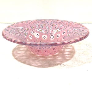 May include: Pink glass bowl with a decorative pattern. The bowl has a wide, shallow shape with a flared rim. The pattern includes white squares, gold dots, and swirling lines. The bowl is likely used for serving or display.