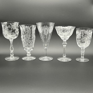 May include: Five clear glass vessels with intricate white etched floral and decorative patterns. The collection includes various shapes, from a tall conical glass to stemmed goblets and a wide-rimmed coupe, all featuring detailed engravings.
