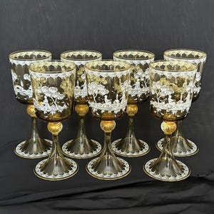 May include: Set of eight amber-colored wine glasses with intricate white and gold painted landscape designs. Each glass features a detailed scene of trees, figures, and architectural elements, with a decorative white rim and gold accents on the stem.