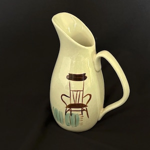 May include: Cream-colored ceramic pitcher with a brown rocking chair design and a light blue fence. The pitcher has a curved handle and a wide spout. A decorative kitchenware item.