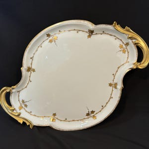 May include: An oval white porcelain serving tray featuring a gold-painted ivy vine pattern along the rim. The tray includes two ornate, curved gold handles on opposite sides, with a smooth, glossy finish and a scalloped edge design.