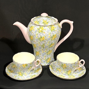 May include: A vintage tea set featuring a teapot and two teacups with saucers. The set has a floral pattern with yellow and blue flowers on a light blue background. The teapot and teacups have pink accents.
