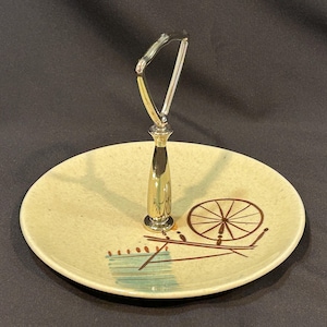 May include: A vintage two-tiered serving tray with a speckled, light yellow plate. The plate features a brown and teal design with a wheel and lines. The handle is silver and gold. A decorative piece for serving food.