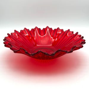 May include: A vibrant red glass bowl featuring a translucent finish and a distinct scalloped, ruffled edge. The interior of the bowl displays a radiating sunburst pattern of embossed lines that extend from the center toward the outer rim.