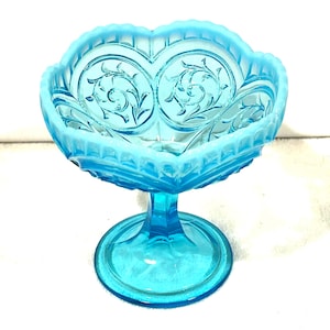 May include: A vintage, light blue glass pedestal candy dish. The dish has a scalloped rim and a decorative floral pattern. The base is round and the stem is a solid column. The glass has a slight opalescent quality.
