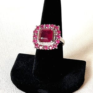 May include: A silver ring featuring a large, square, deep red gemstone at the center, surrounded by smaller, round red gemstones. The ring is displayed on a black velvet ring holder.