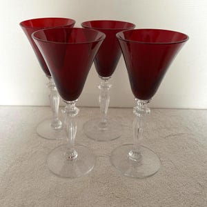 May include: Four ruby red wine glasses with clear stems and bases. The glasses have a classic design with a flared bowl and a long, elegant stem. The glasses are arranged in a group, showcasing their color and shape.
