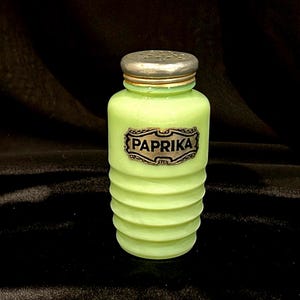 May include: A vintage, light green glass paprika shaker with a silver-colored metal lid. The shaker has a ribbed design and a label that reads "PAPRIKA" in a decorative font. The background is black.
