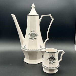 May include: A tall, off-white ceramic coffee pot with a pointed lid and a matching smaller creamer pitcher. Both pieces feature a hexagonal shape, a ribbed base, and a dark gray floral pattern with a scalloped border detail on the main body.