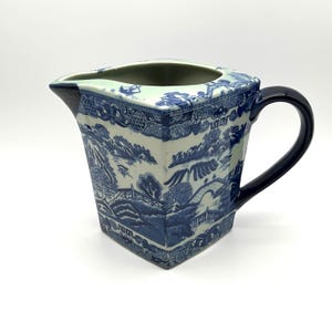May include: A square-shaped ceramic creamer pitcher featuring a blue and white transferware pattern depicting a landscape with bridges, trees, and pagodas. The pitcher has a dark blue handle and a wide pouring spout with a geometric border design.