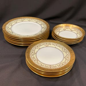 May include: A set of white ceramic dinnerware featuring a wide gold rim with an intricate filigree pattern. The collection includes a stack of large dinner plates, a stack of smaller salad plates, and a stack of shallow bowls with gold trim.