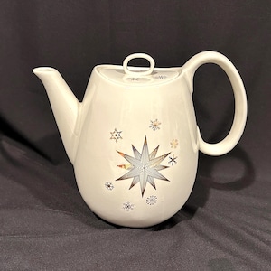 May include: Cream-colored ceramic teapot with a lid and a large handle. The teapot is decorated with a large starburst design and smaller snowflake patterns in gold and black. The spout is long and slender.