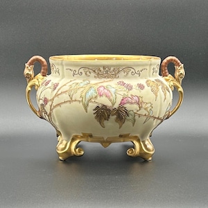 May include: A cream-colored ceramic bowl with two ornate gold handles and three gold feet. The exterior features a painted design of brown, pink, and green bramble vines with berries, accented by gold filigree patterns along the upper rim.