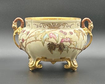 May include: A cream-coloured ceramic bowl with two ornate gold handles and three gold feet. The exterior features a painted design of brown, pink, and green bramble vines with berries, accented by gold filigree patterns along the upper rim.