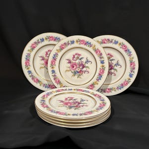 May include: A set of vintage porcelain plates with a floral design. The plates feature a cream-colored background with a central bouquet of pink, purple, and blue flowers. The rims are adorned with a border of pink roses, blue and yellow accents, and gold trim.