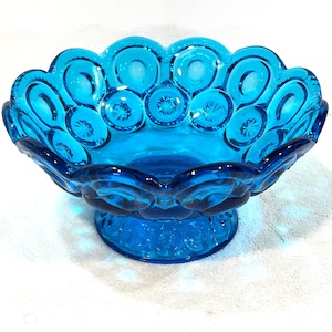May include: A vibrant blue glass bowl with a pedestal base. The bowl features a scalloped rim with a repeating pattern of circles and starbursts. The bowl is made of a clear, transparent glass.