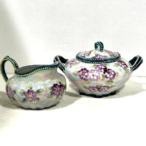 May include: A hand-painted ceramic tea set featuring a creamer and a covered teapot. The set is decorated with purple flowers and green accents on a light blue background. The handles and rims are detailed with a dark green trim.