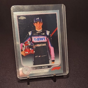Puede incluir: Tarjeta coleccionable de Esteban Ocon con traje de carreras, sosteniendo un casco. La tarjeta está en una funda protectora transparente y sobre un pequeño soporte. Presenta el logotipo del equipo Alpine F1.