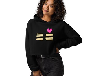 May include: Black cropped hoodie with a pink heart and two sets of horizontal gold lines. The hoodie has a drawstring hood and long sleeves. The model is wearing black jeans and a black belt.