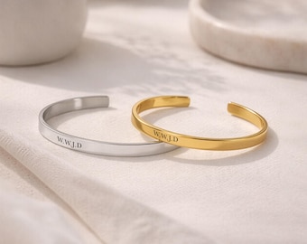 May include: Two open cuff bracelets, one in a silver tone and one in a gold tone, each featuring a smooth, polished finish and the engraved text W.W.J.D. on the outer surface. These minimalist metal bangles have a simple, thin, and open design.
