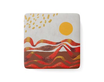 May include: A square coaster featuring a white background with a yellow sun and gold dash patterns. The lower portion displays layered, wavy horizontal bands in shades of red, orange, and brown, creating a textured landscape design.