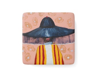 May include: A square ceramic coaster featuring a painted illustration of a figure wearing a wide-brimmed dark blue hat, a white collar, and a yellow and red striped garment against a light pink background decorated with faint, circular floral patterns.