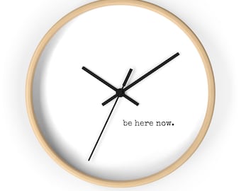 May include: A round wall clock with a light wood frame and a white face. The clock features black hands and the phrase "be here now" in a simple, black font. The clock's design is minimalist and modern.