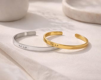 May include: Two open cuff bracelets, one in a silver tone and one in a gold tone, each featuring the engraved text S.C.A.P. on the outer surface. These thin, smooth metal bangles have a simple, circular design with an opening at the back.
